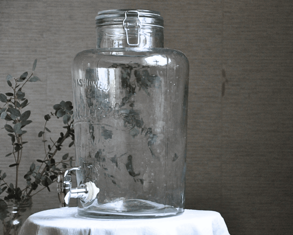 Glasbehållare med tappkran på ett bord med vit duk.